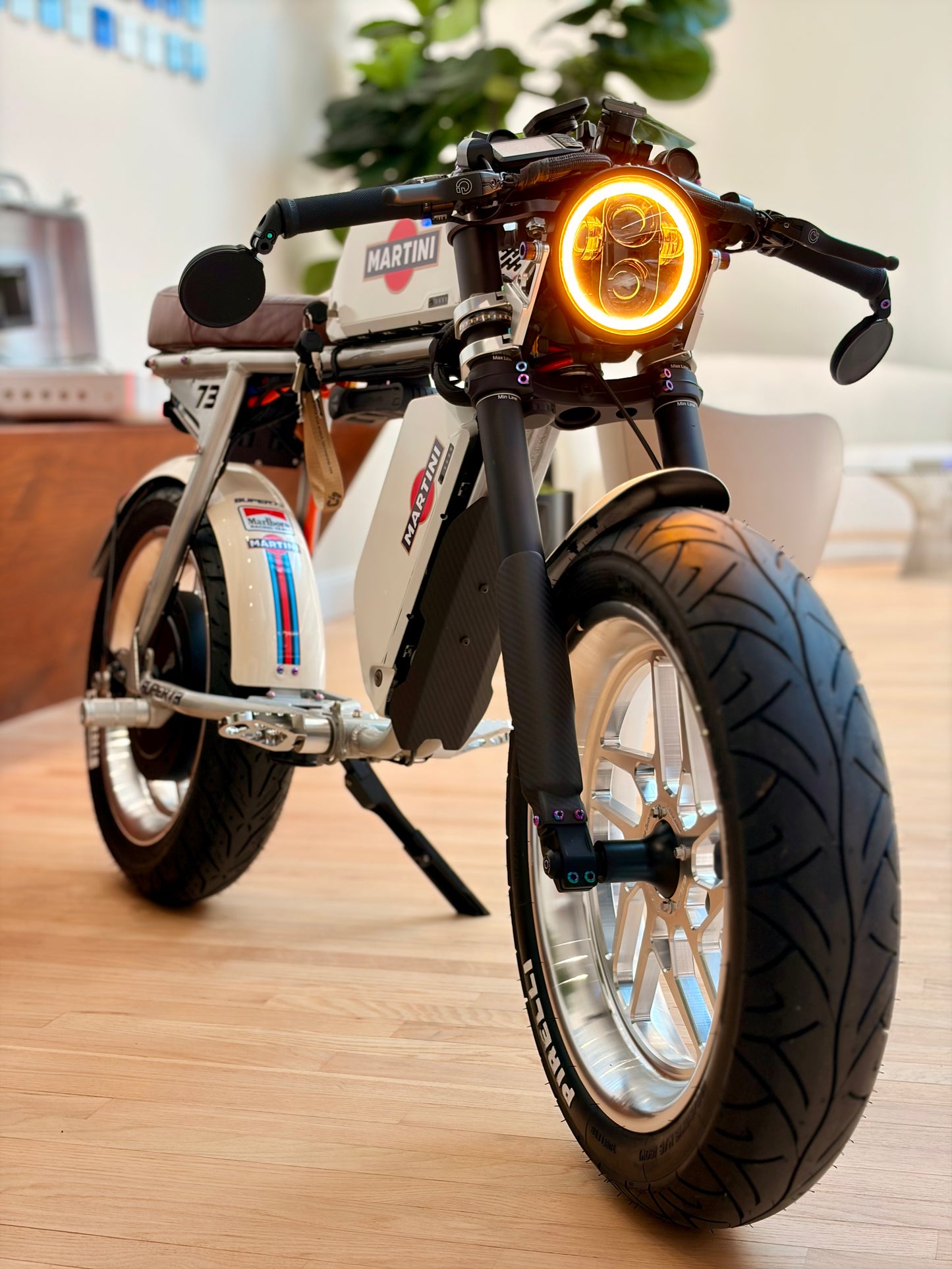 A retro-style electric motorcycle with a white frame, black forks and tires, and a glowing amber headlight, photographed indoors on a light wood floor.