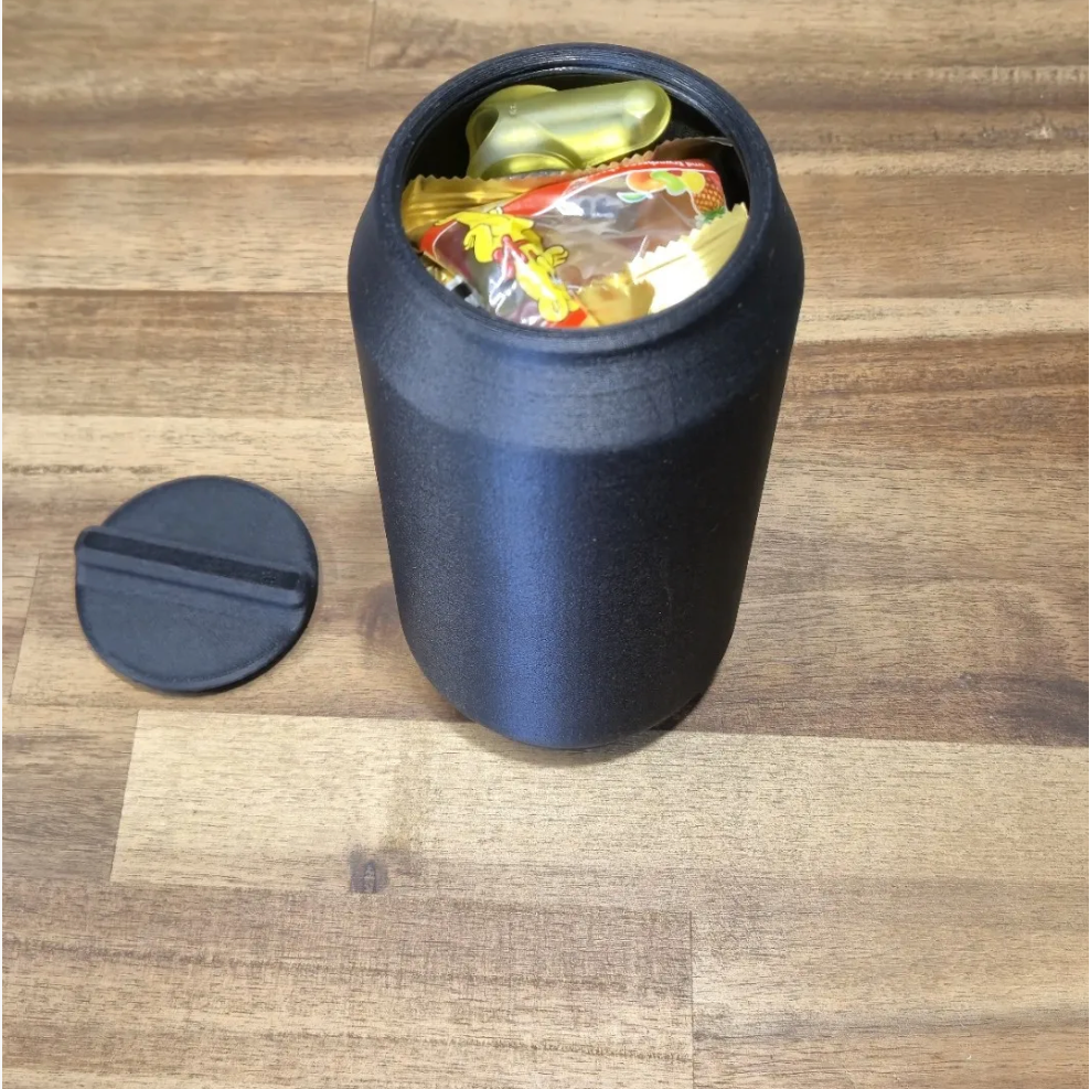 A matte black cylindrical storage container with its lid off, showing a gold capsule-shaped item and colorful wrapped candies inside, placed on a light brown wood-grain surface.