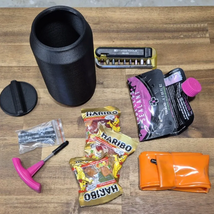 A flat-lay arrangement of various emergency preparedness and survival tools on a light brown wooden surface, including a black insulated water bottle, a black cap, a pink T-handle tool, a Haribo candy pack, a black and pink first aid pouch, a metal tool, and an orange pouch.