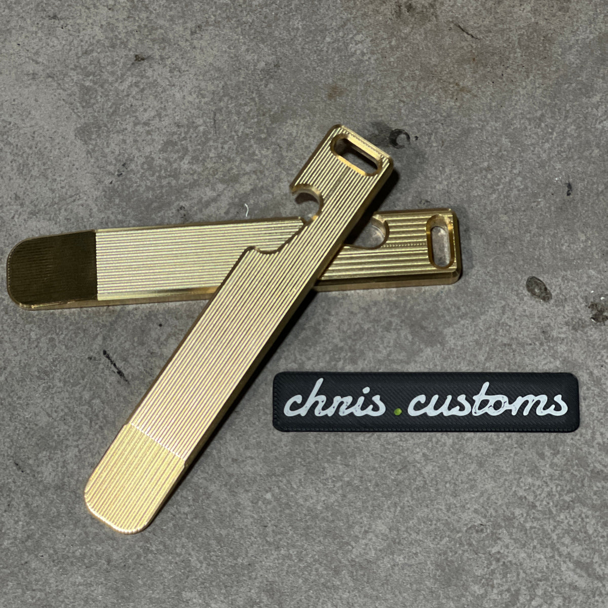 A gold-colored metal multi-tool keychain with a textured ribbed finish, featuring a bottle opener, small knife blade, and pocket clip. The keychain is displayed on a gray concrete surface next to a black custom license plate frame with the text 'chris.
