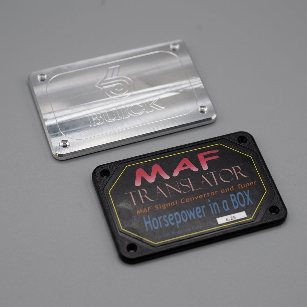 Two automotive badges or covers on a white sheet of paper: a clear engraved rectangular plate labeled 'BUICK' and a black rounded-rectangle plate labeled 'MAF TRANSLATOR Horsepower in a BOX' with red, blue, and yellow text, placed on a light wood surface.