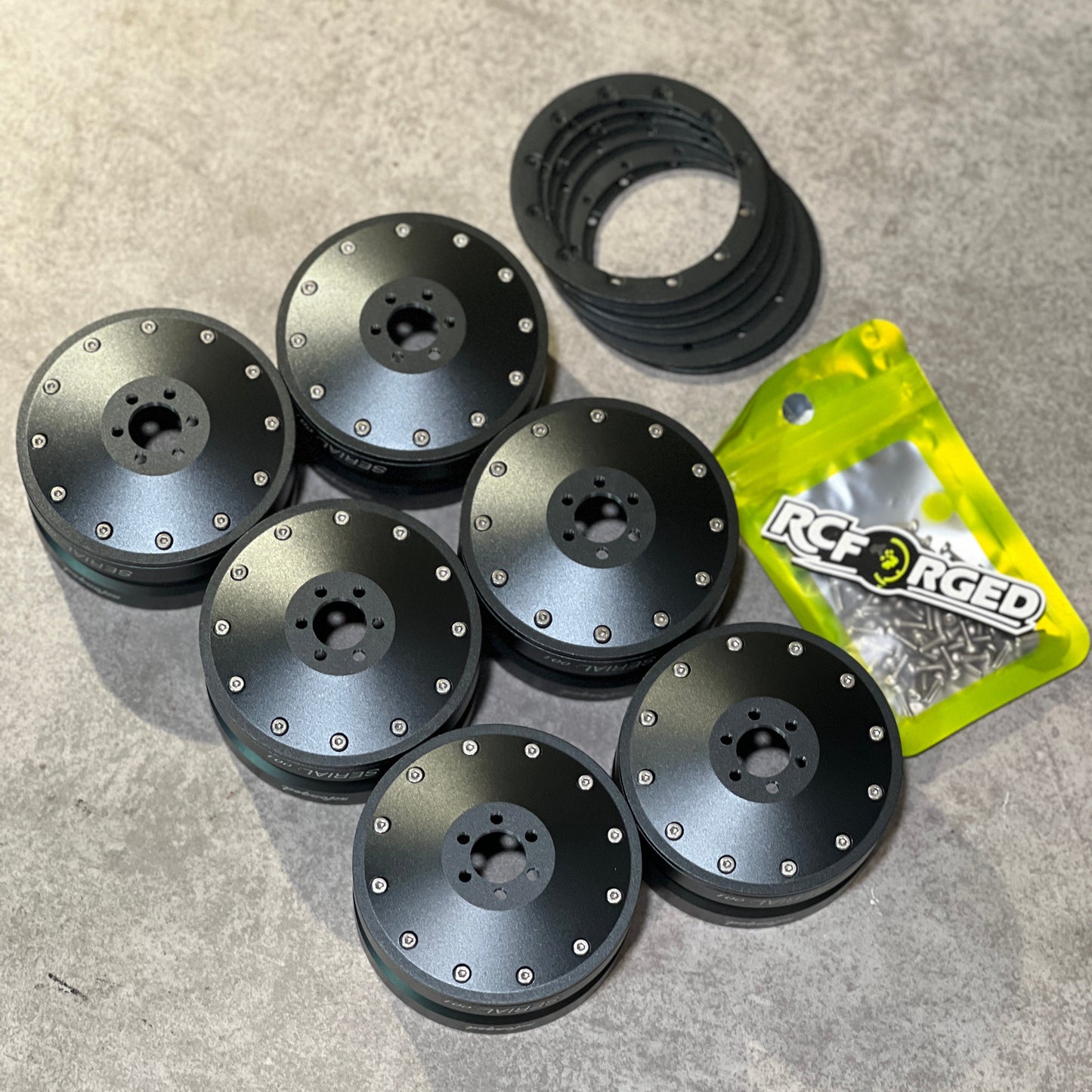 Six black brushed metal RC car wheel spacers with silver rivets, a black rubber ring, and a yellow bag of RC Racing brand screws on a gray concrete floor.