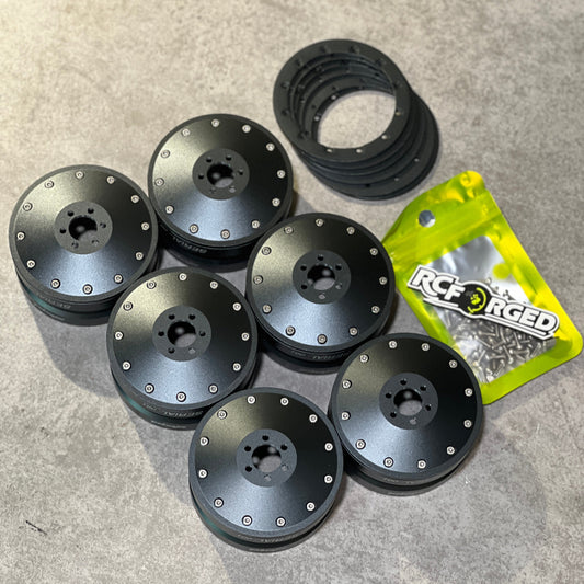 Six black brushed metal RC car wheel spacers with silver rivets, a black rubber ring, and a yellow bag of RC Racing brand screws on a gray concrete floor.