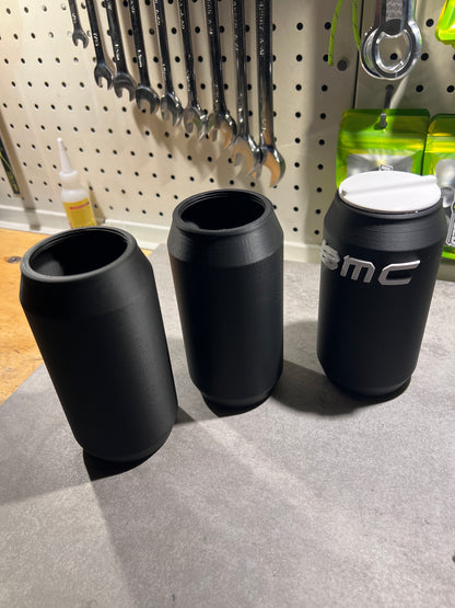 Three matte black cylindrical cups or tumbler-like containers arranged on a light gray surface, with a pegboard and tools visible in the background. One cup has a white lid and a partial white logo reading "MC".