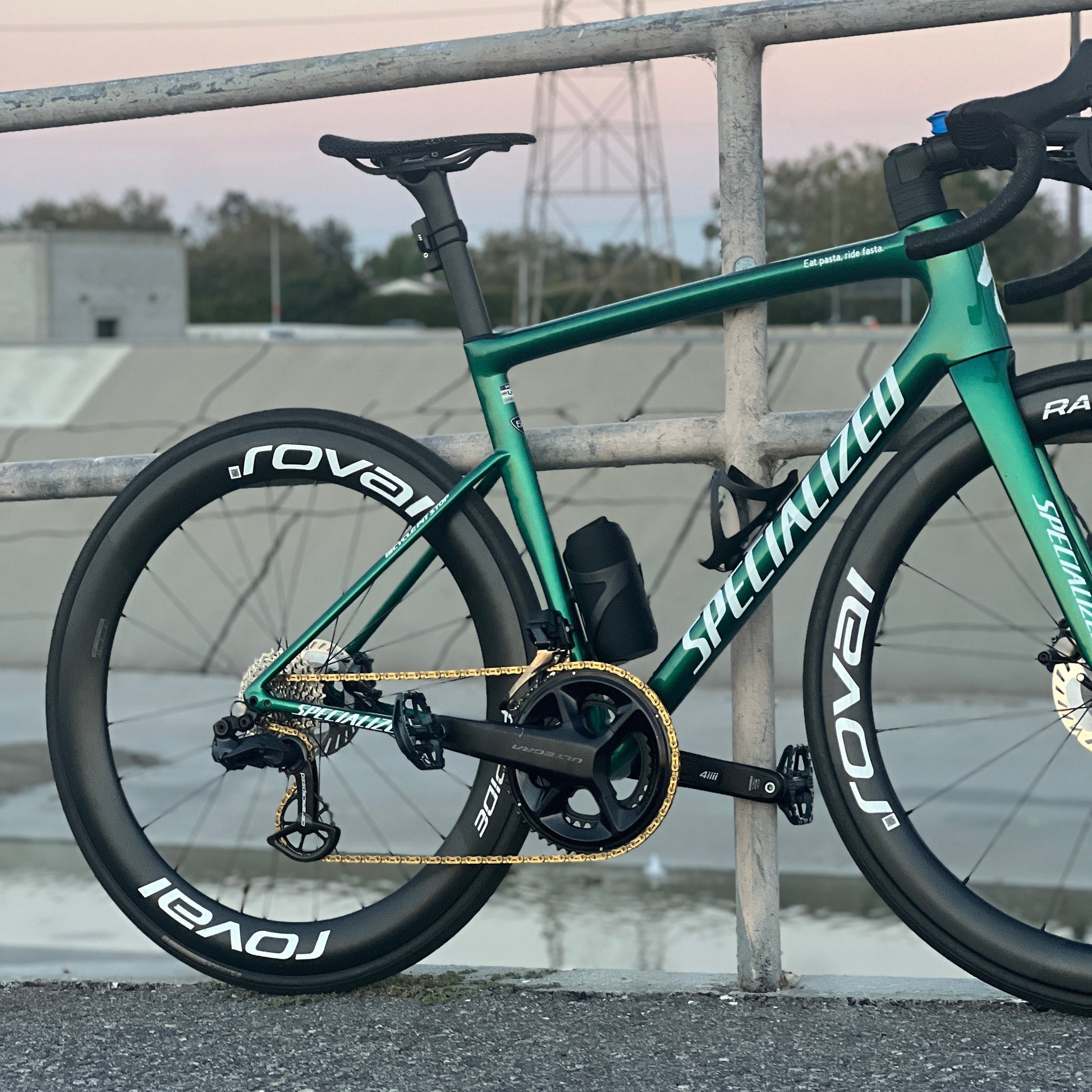 A green Specialized road bicycle with black Roval wheels and a gold chain, leaning against a metal railing in an outdoor setting.