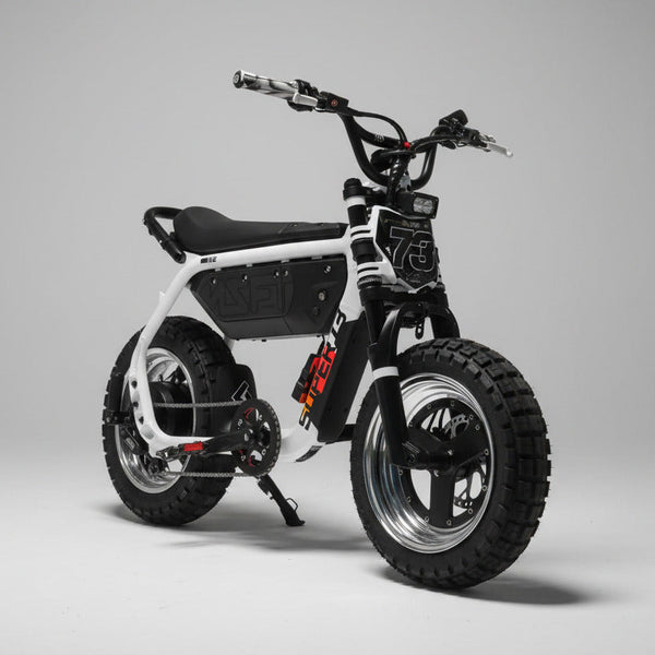 A white and black electric dirt bike with knobby tires, a black seat, and a battery mounted on the frame, parked on an asphalt surface outdoors with trees and a blue sky in the background.