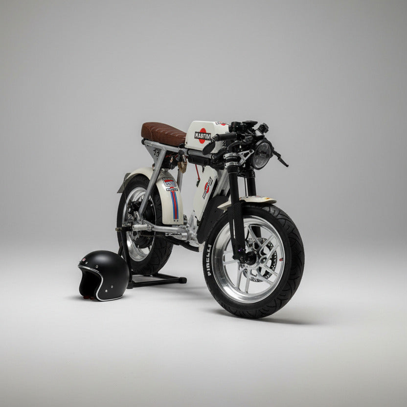 A custom-built, low-profile electric motorcycle with a white body, black tires, and a brown leather seat, parked on a light wood floor in a showroom. A black helmet and a silver electronic device sit on a wooden bench in the background.