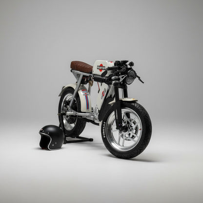 A custom-built, low-profile electric motorcycle with a white body, black tires, and a brown leather seat, parked on a light wood floor in a showroom. A black helmet and a silver electronic device sit on a wooden bench in the background.