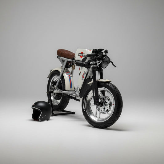 A custom-built, low-profile electric motorcycle with a white body, black tires, and a brown leather seat, parked on a light wood floor in a showroom. A black helmet and a silver electronic device sit on a wooden bench in the background.