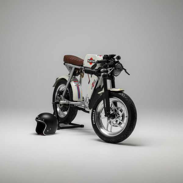 A custom-built, low-profile electric motorcycle with a white body, black tires, and a brown leather seat, parked on a light wood floor in a showroom. A black helmet and a silver electronic device sit on a wooden bench in the background.