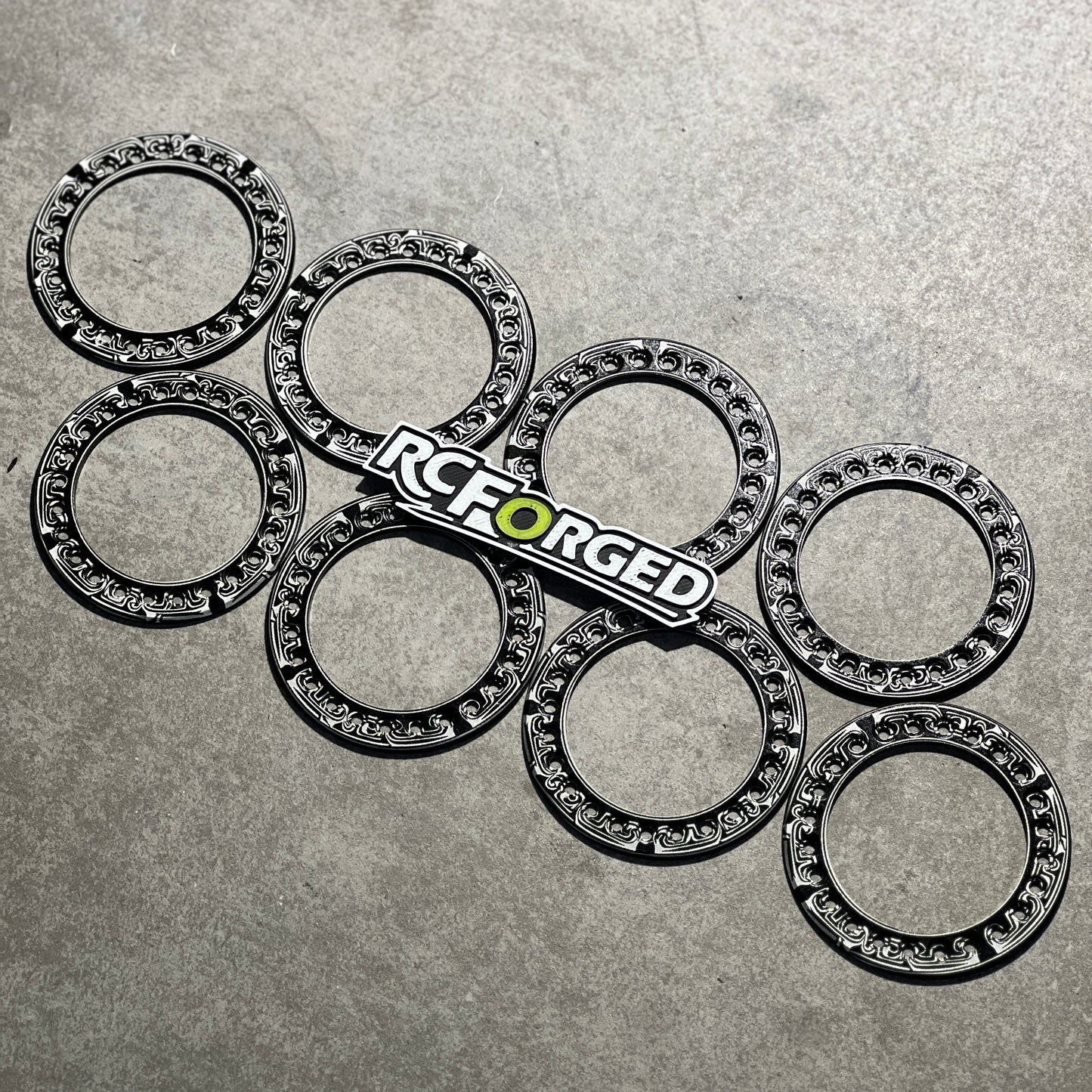 Six black metal caster wheels with silver ball bearings inside, arranged on a textured gray surface, with an RC Forged logo sticker placed in the center.
