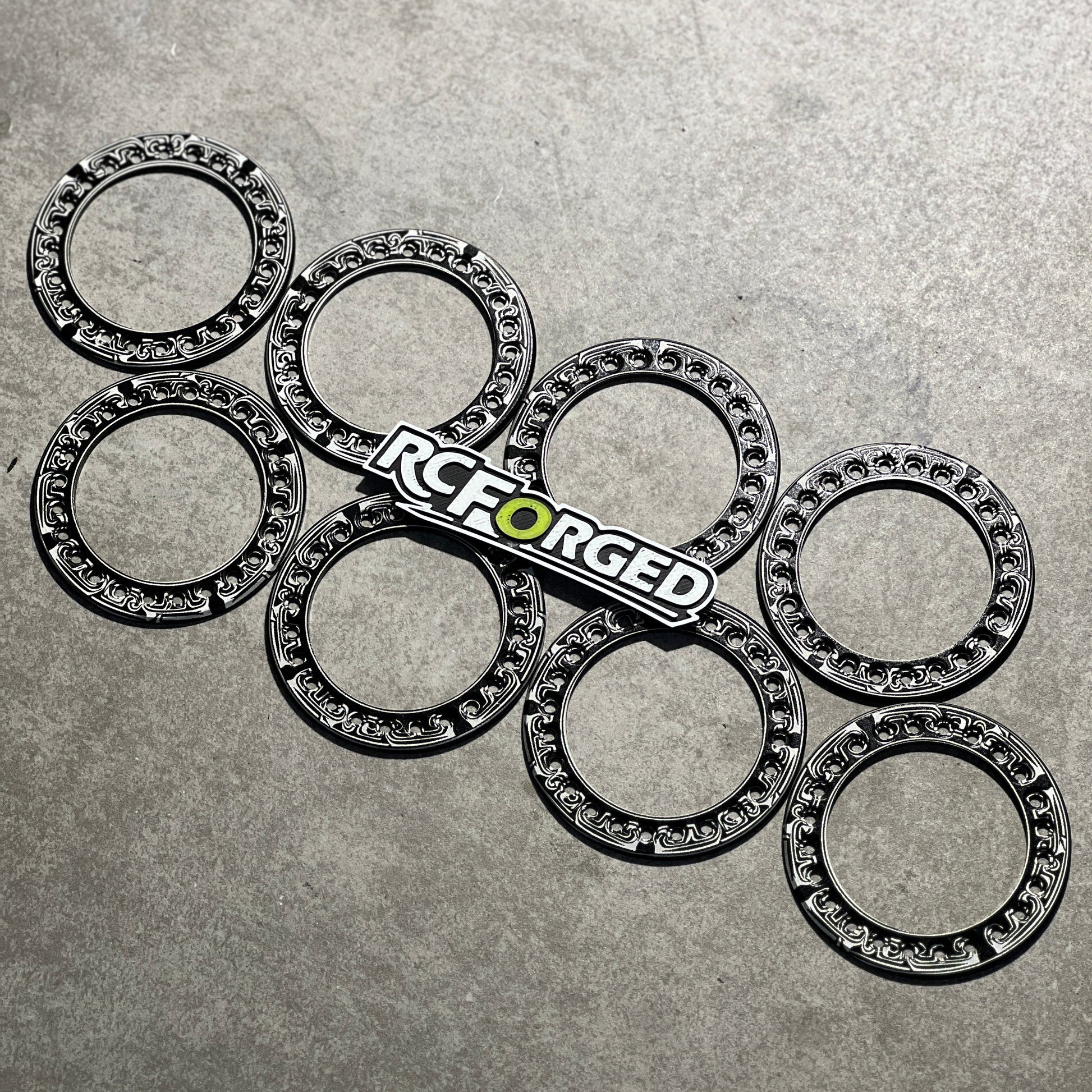 Six black metal caster wheels with silver ball bearings inside, arranged on a textured gray surface, with an RC Forged logo sticker placed in the center.