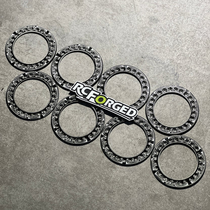 Six black metal caster wheels with silver ball bearings inside, arranged on a textured gray surface, with an RC Forged logo sticker placed in the center.