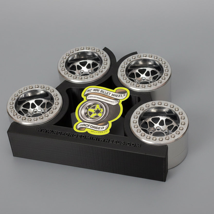 A set of gray anodized finish beadlock wheels for remote control trucks, displayed on a black stand with measuring grid background.