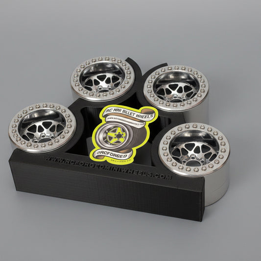 A set of gray anodized finish beadlock wheels for remote control trucks, displayed on a black stand with measuring grid background.