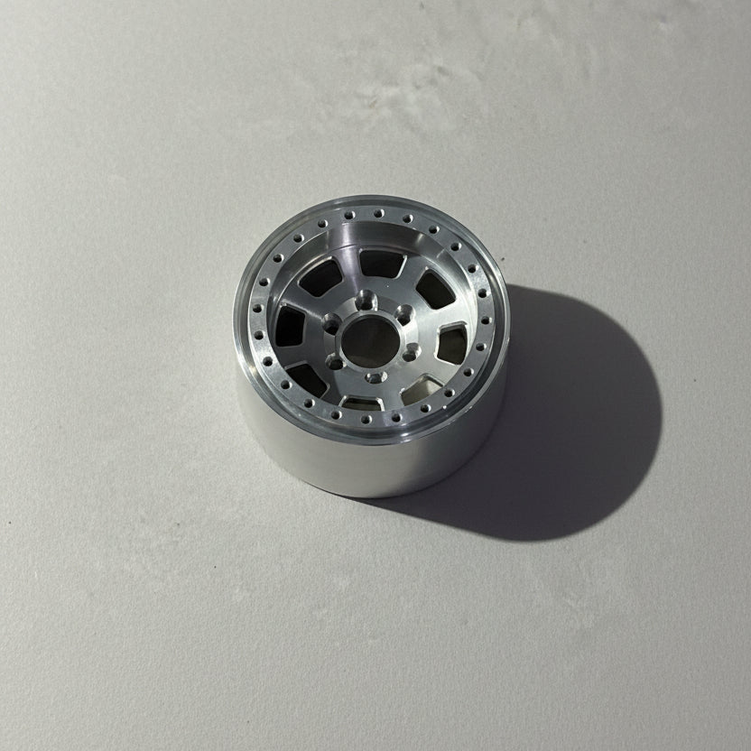 A silver metallic multi-spoke wheel rim with a drilled outer edge and center bore, resting on a textured gray concrete floor.