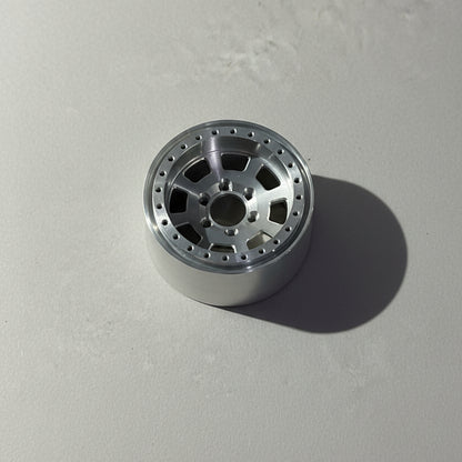 A silver metallic multi-spoke wheel rim with a drilled outer edge and center bore, resting on a textured gray concrete floor.