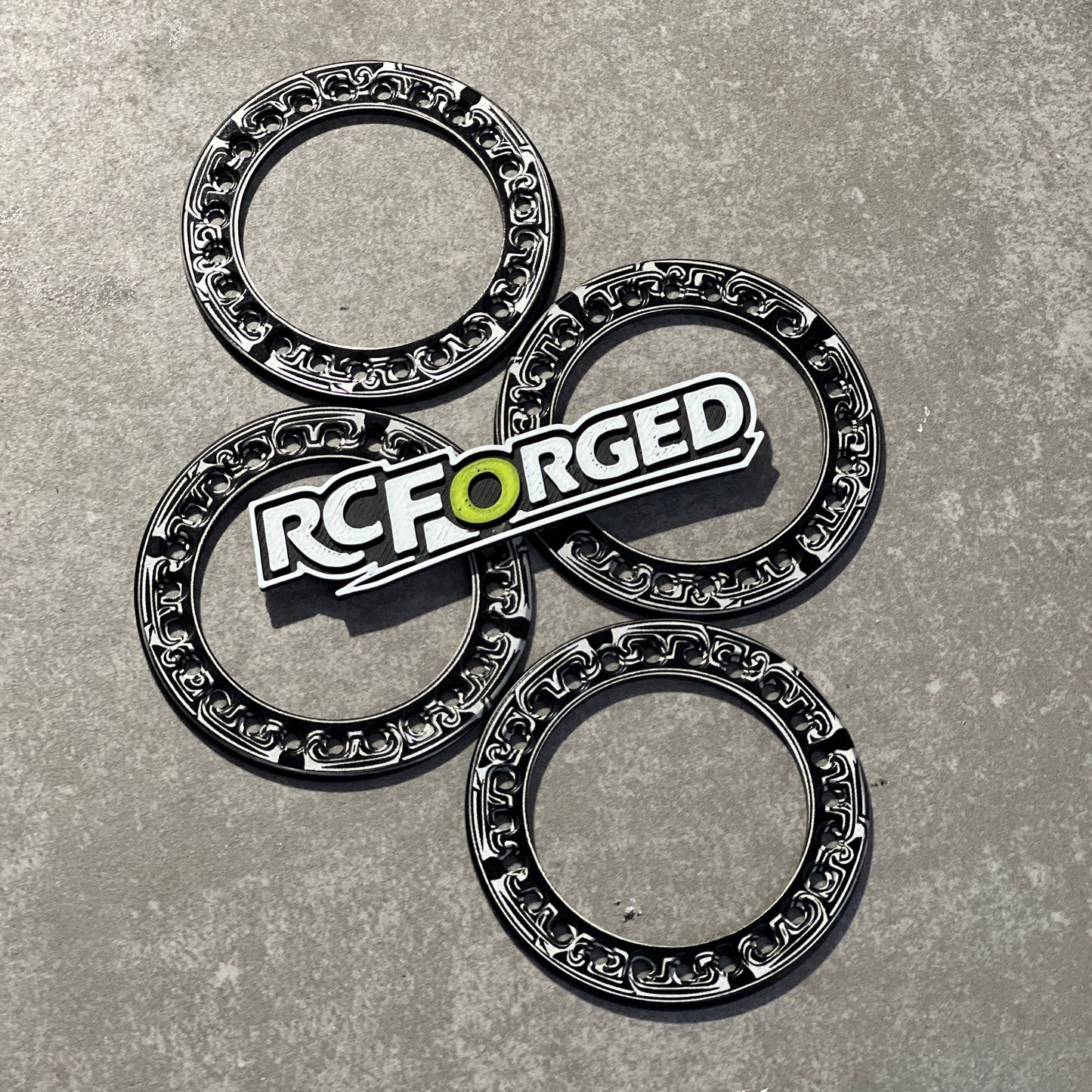 Four black and silver metallic ring-shaped components arranged on a gray textured surface, with a prominent RC FORGED logo badge placed over the center piece.