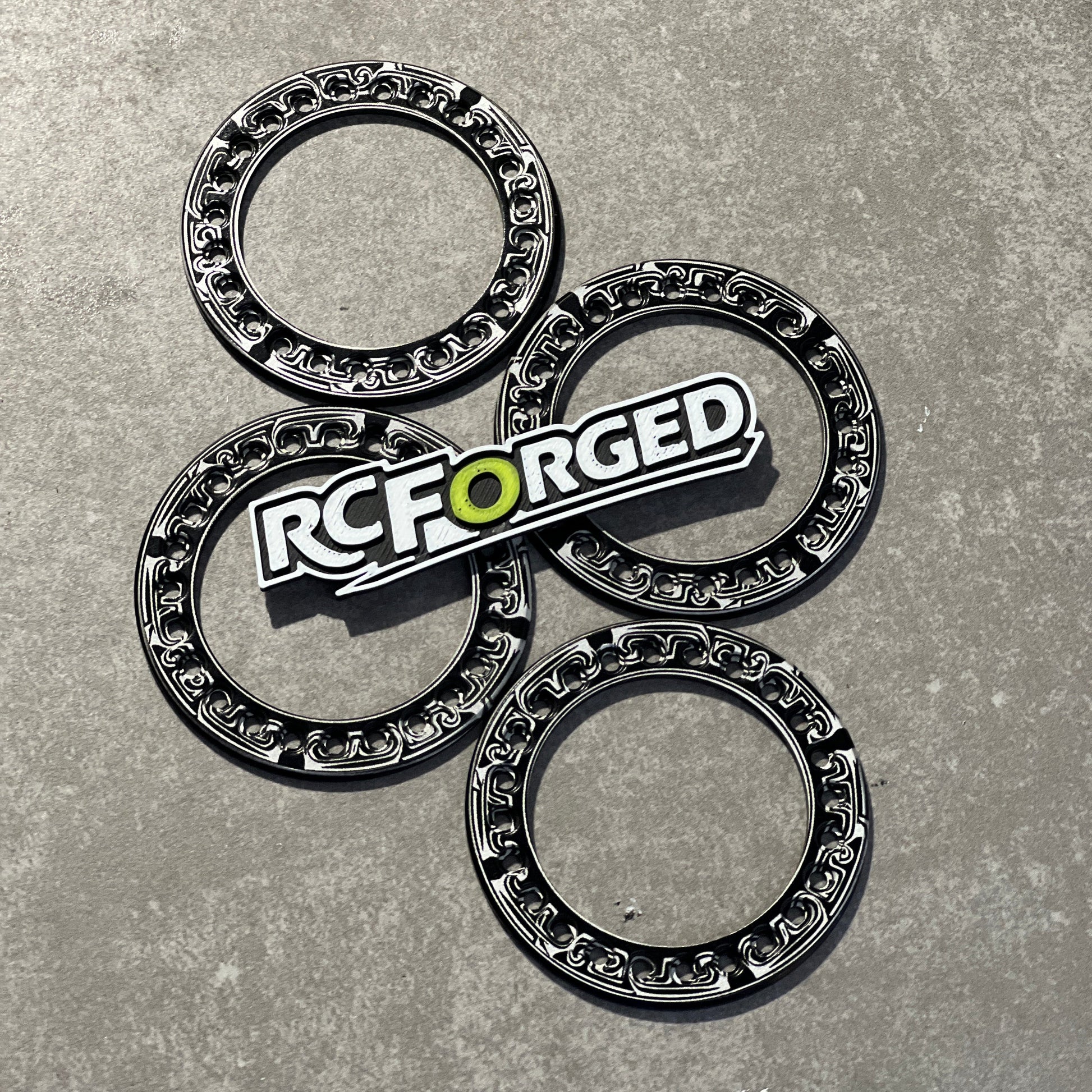 Four black and silver metallic ring-shaped components arranged on a gray textured surface, with a prominent RC FORGED logo badge placed over the center piece.