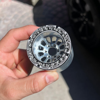 A hand holding a chrome silver decorative wheel rim with an ornate black outer border, set against a blurred outdoor background with a car wheel and pavement.