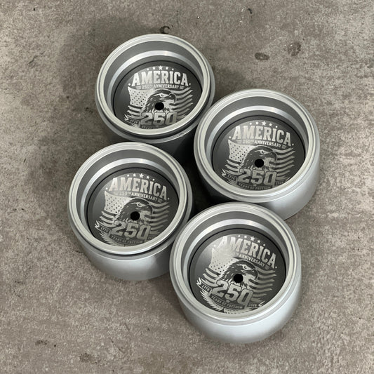 Four silver metal yo-yos arranged in a square on a gray concrete floor. Each yo-yo features a custom circular center label with the text 'AMERICA YOUTUBING 250' and an eagle graphic.
