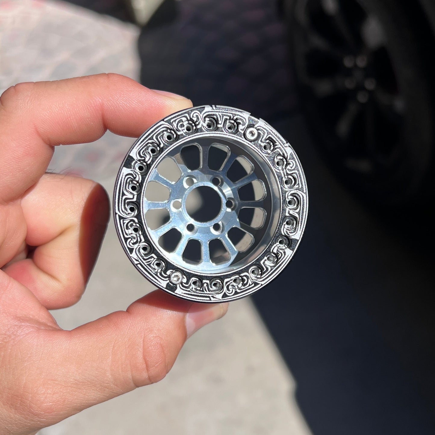 A hand holding a small silver metal wheel-shaped accessory with an ornate patterned outer rim, photographed outdoors near a black car wheel.