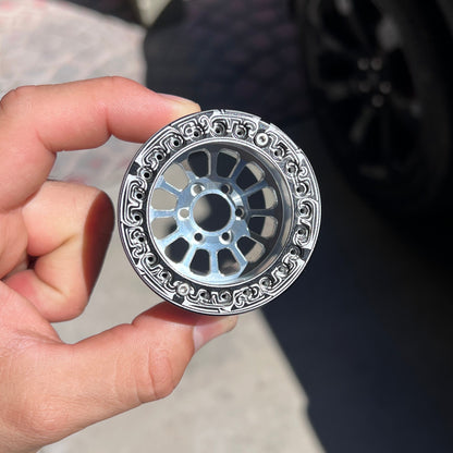 A hand holding a small silver metal wheel-shaped accessory with an ornate patterned outer rim, photographed outdoors near a black car wheel.